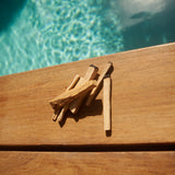 6 sticks of palo santo sitting on a pool deck in a hotel in Tulum.  Each stick is a different thickness and cut differently.  Some are wildly cut and some are straight cut.  The water of the pool which peeks through is aquamarine in colour.