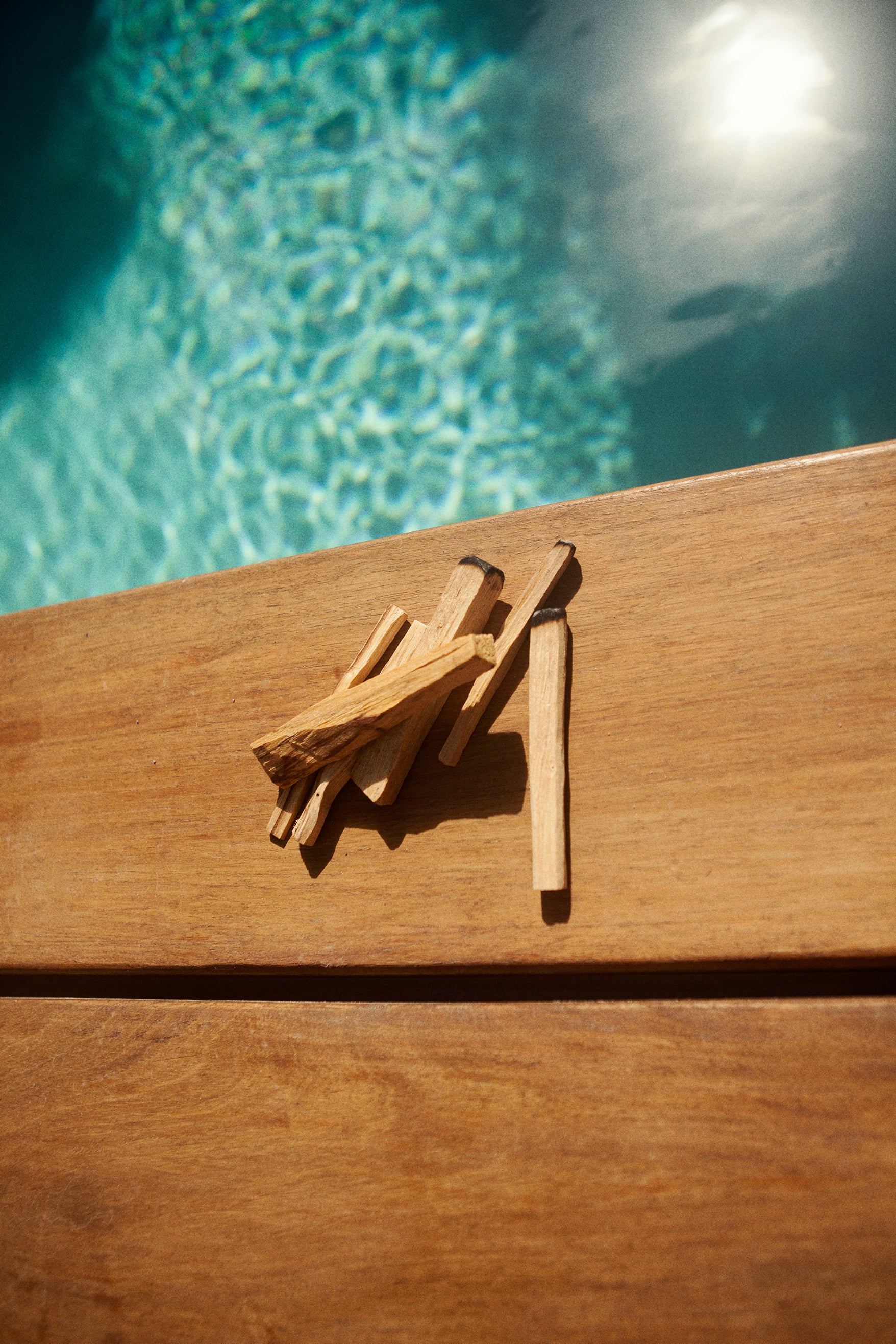 6 sticks of palo santo sitting on a pool deck in a hotel in Tulum.  Each stick is a different thickness and cut differently.  Some are wildly cut and some are straight cut.  The water of the pool which peeks through is aquamarine in colour.