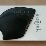 a palm sized black guasha tool (made from volcanic and metero matter) shaped into an abstract shape with one side ridged. The brand logo is engraved in a metallic copper colour.  The tool sitting on the drawer box it is sent in.