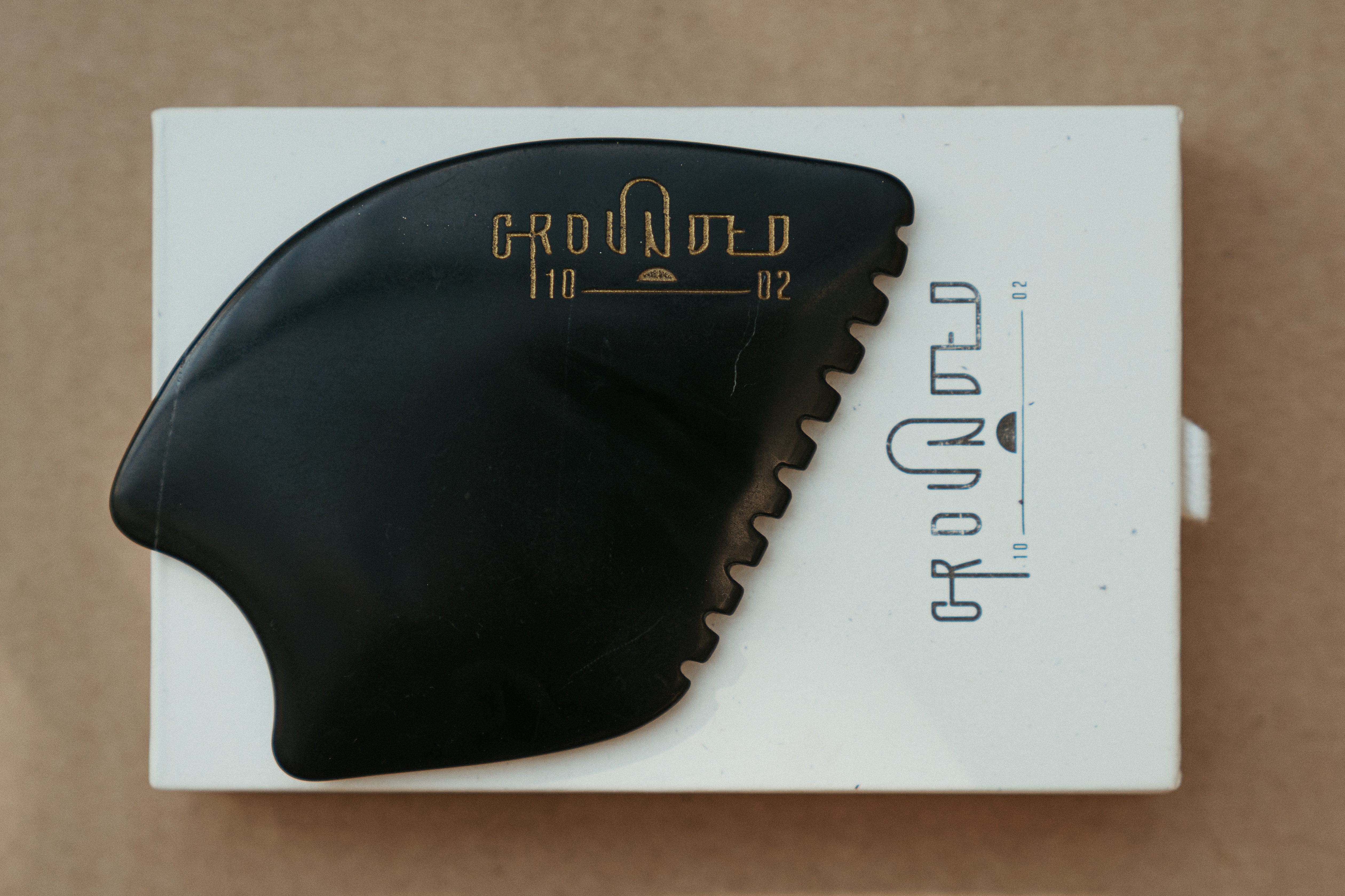 a palm sized black guasha tool (made from volcanic and metero matter) shaped into an abstract shape with one side ridged. The brand logo is engraved in a metallic copper colour.  The tool sitting on the drawer box it is sent in.
