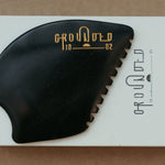 a palm sized black guasha tool (made from volcanic and metero matter) shaped into an abstract shape with one side ridged. The brand logo is engraved in a metallic bright yellow gold colour.  The tool sitting on the drawer box it is sent in.