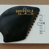 a palm sized black guasha tool (made from volcanic and metero matter) shaped into an abstract shape with one side ridged. The brand logo is engraved in a metallic bright yellow gold colour.  The tool sitting on the drawer box it is sent in.