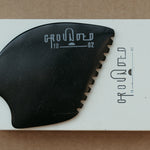 a palm sized black guasha tool (made from volcanic and metero matter) shaped into an abstract shape with one side ridged. The brand logo is engraved in a matt grey colour.  The tool sitting on the drawer box it is sent in.