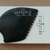 a palm sized black guasha tool (made from volcanic and metero matter) shaped into an abstract shape with one side ridged. The brand logo is engraved in a matt grey colour.  The tool sitting on the drawer box it is sent in.