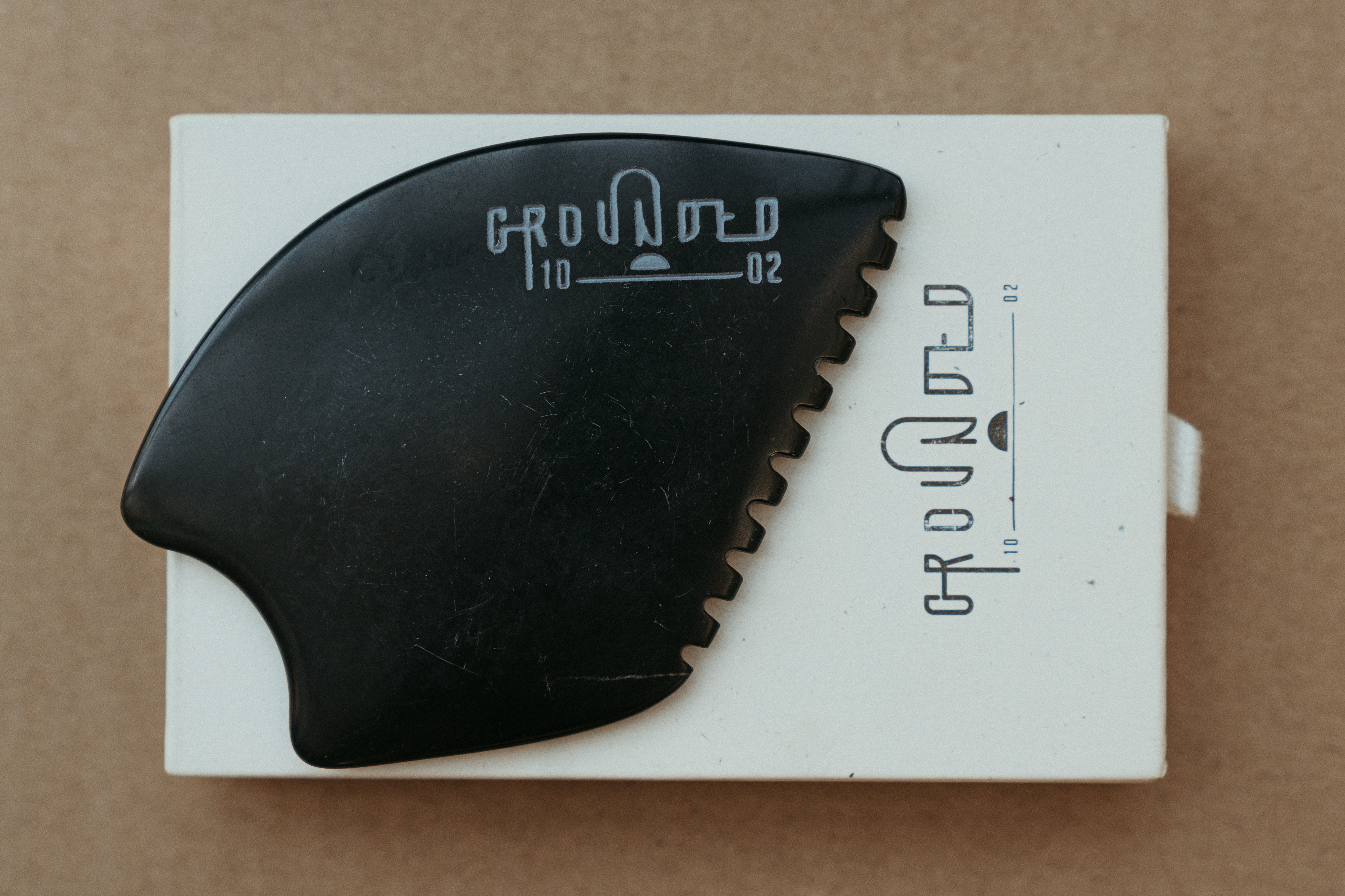 a palm sized black guasha tool (made from volcanic and metero matter) shaped into an abstract shape with one side ridged. The brand logo is engraved in a matt grey colour.  The tool sitting on the drawer box it is sent in.