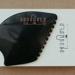 a palm sized black guasha tool (made from volcanic and metero matter) shaped into an abstract shape with one side ridged. The brand logo is engraved in a metallic rose gold  colour.  The tool sitting on the drawer box it is sent in.