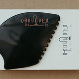 a palm sized black guasha tool (made from volcanic and metero matter) shaped into an abstract shape with one side ridged. The brand logo is engraved in a metallic rose gold  colour.  The tool sitting on the drawer box it is sent in.
