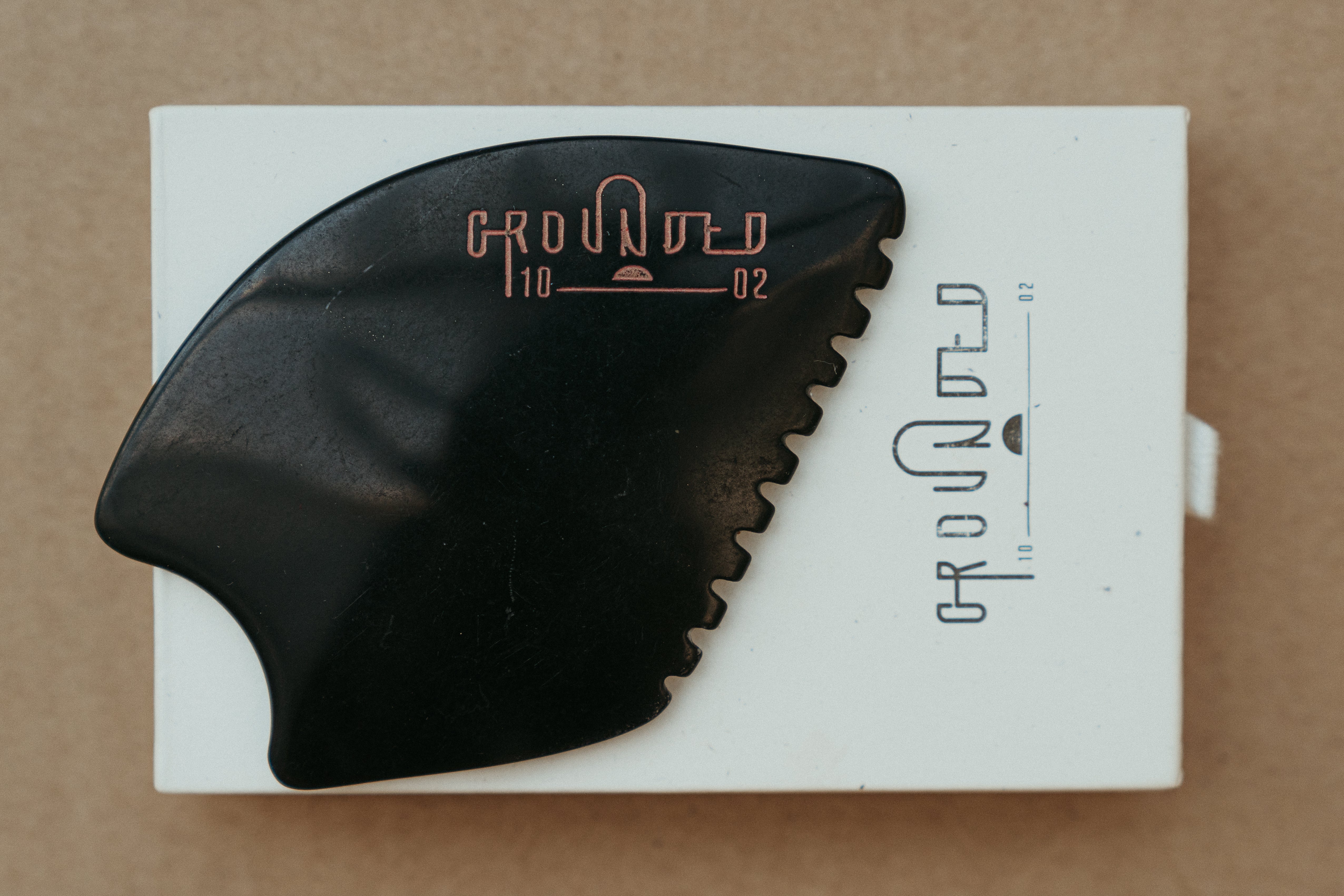 a palm sized black guasha tool (made from volcanic and metero matter) shaped into an abstract shape with one side ridged. The brand logo is engraved in a metallic rose gold  colour.  The tool sitting on the drawer box it is sent in.