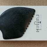 a palm sized black guasha tool (made from volcanic and metero matter) shaped into an abstract shape with one side ridged. The brand logo is engraved with no colour (just etched).  The tool sitting on the drawer box it is sent in.