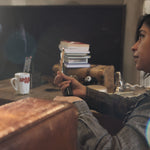 A lady sitting in a messy family room holding a very thin stick of Palo Santo with a light flame on top.  It looks like she is in a serious conversation.