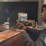 A lady sitting in a messy family room holding a very thin stick of Palo Santo with a light flame on top.  It looks like she is in a serious conversation.