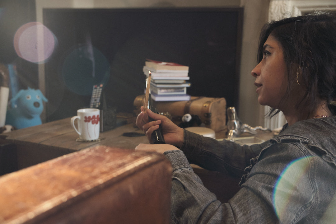 A lady sitting in a messy family room holding a very thin stick of Palo Santo with a light flame on top.  It looks like she is in a serious conversation.