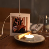 A small poleroid picture is standing on a restaurant table in Notting Hill.  the picture is of a lady wearing gloves and holding a #rockwithcare stone (a guasha).  In front of the picture, on a small tea plate, is a lit tea light candle and a palo santo stick.