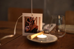 A small poleroid picture is standing on a restaurant table in Notting Hill.  the picture is of a lady wearing gloves and holding a #rockwithcare stone (a guasha).  In front of the picture, on a small tea plate, is a lit tea light candle and a palo santo stick.