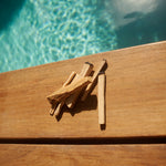 6 sticks of palo santo sitting on a pool deck in a hotel in Tulum.  Each stick is a different thickness and cut differently.  Some are wildly cut and some are straight cut.  The water of the pool which peeks through is aquamarine in colour.
