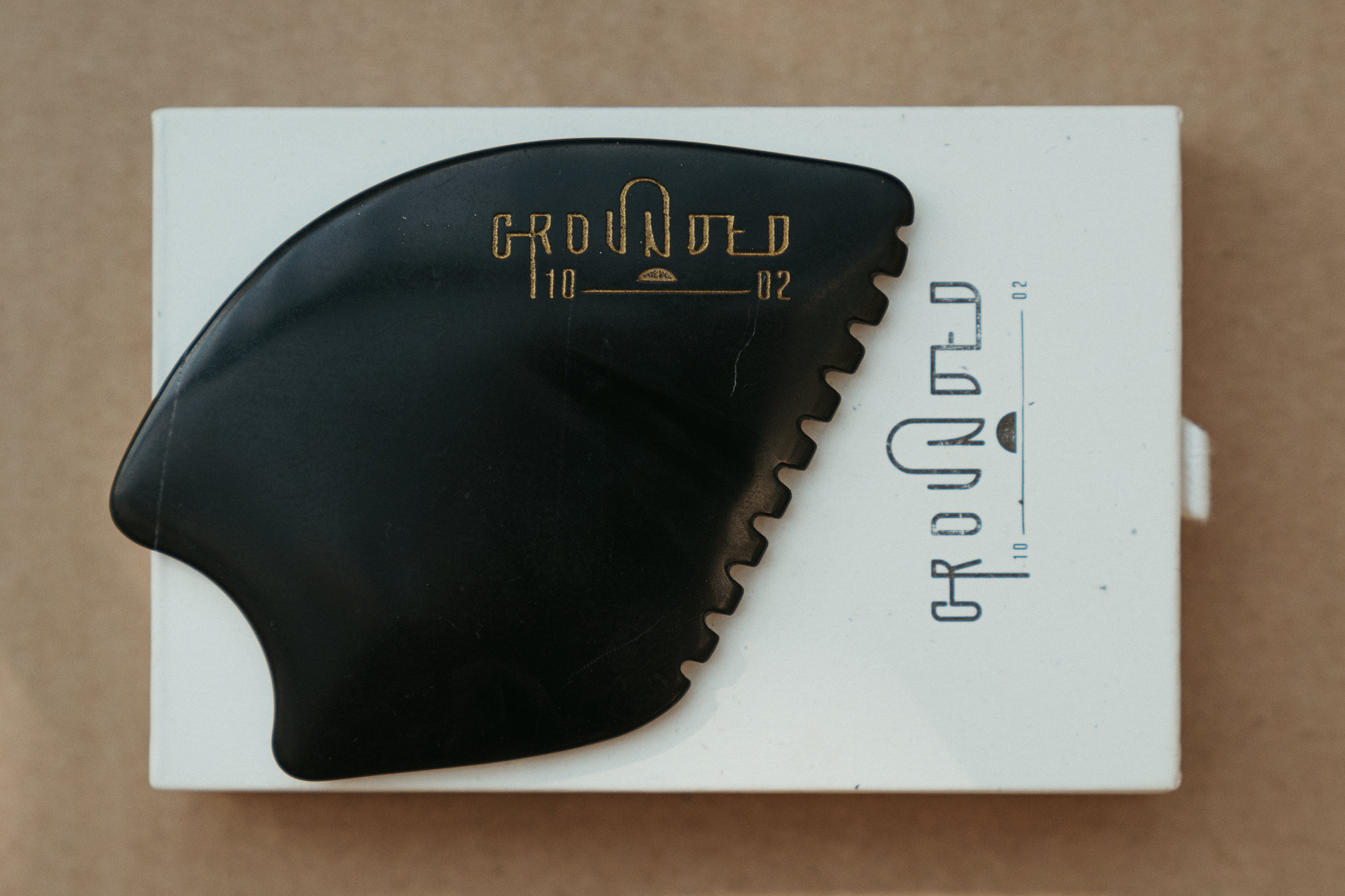 a palm sized black guasha tool (made from volcanic and metero matter) shaped into an abstract shape with one side ridged. The brand logo is engraved in a metallic copper colour.  The tool sitting on the drawer box it is sent in. 