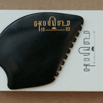 a palm sized black guasha tool (made from volcanic and metero matter) shaped into an abstract shape with one side ridged. The brand logo is engraved in a metallic bright yellow gold colour.  The tool sitting on the drawer box it is sent in. 