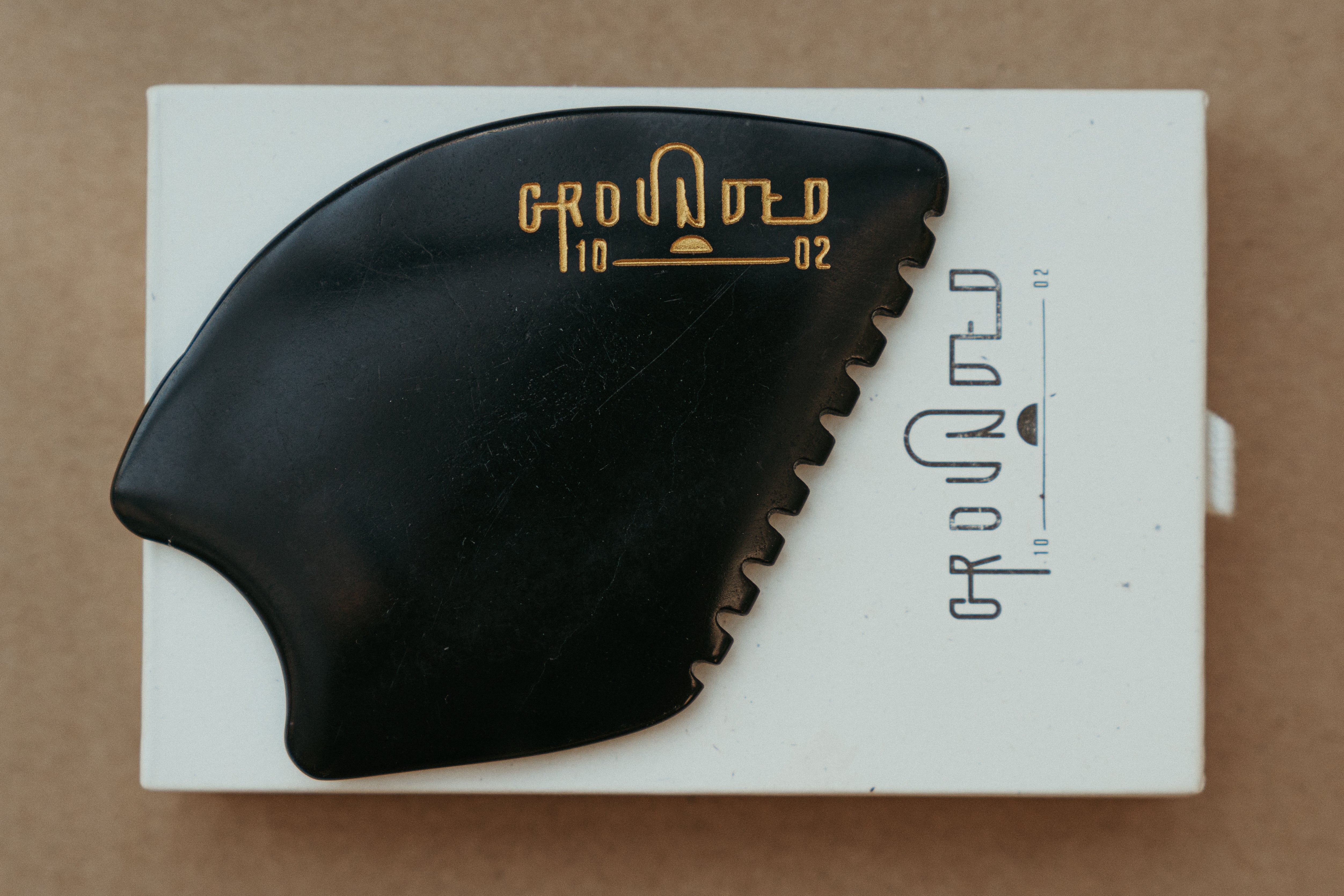 a palm sized black guasha tool (made from volcanic and metero matter) shaped into an abstract shape with one side ridged. The brand logo is engraved in a metallic bright yellow gold colour.  The tool sitting on the drawer box it is sent in. 