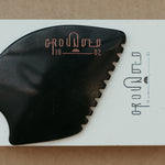 a palm sized black guasha tool (made from volcanic and metero matter) shaped into an abstract shape with one side ridged. The brand logo is engraved in a metallic rose gold  colour.  The tool sitting on the drawer box it is sent in. 