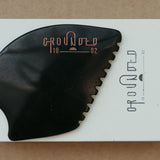 a palm sized black guasha tool (made from volcanic and metero matter) shaped into an abstract shape with one side ridged. The brand logo is engraved in a metallic rose gold  colour.  The tool sitting on the drawer box it is sent in. 