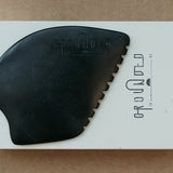 a palm sized black guasha tool (made from volcanic and metero matter) shaped into an abstract shape with one side ridged. The brand logo is engraved with no colour (just etched).  The tool sitting on the drawer box it is sent in. 