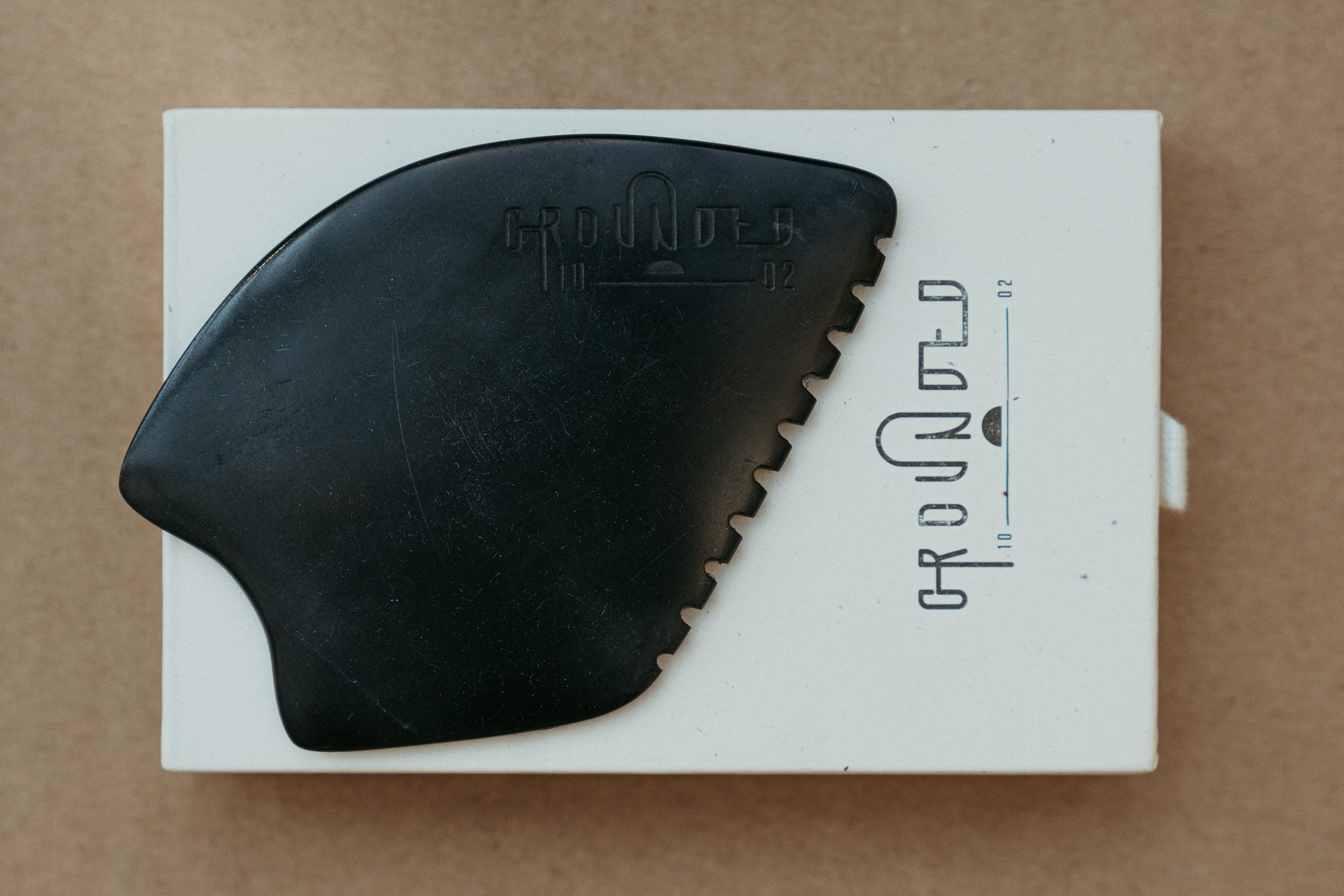 a palm sized black guasha tool (made from volcanic and metero matter) shaped into an abstract shape with one side ridged. The brand logo is engraved with no colour (just etched).  The tool sitting on the drawer box it is sent in. 