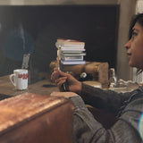 A lady sitting in a messy family room holding a very thin stick of Palo Santo with a light flame on top.  It looks like she is in a serious conversation. 